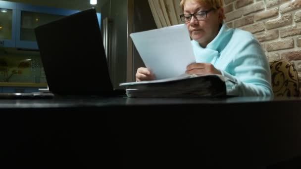 Oudere vrouw die thuis met de documenten werkt met een laptop. — Stockvideo