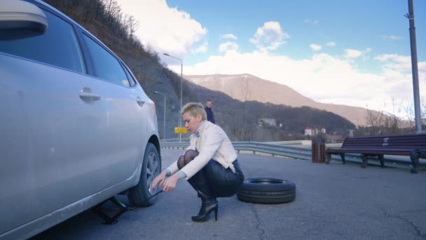 Humor. mulher mudando uma roda de carro. um homem cavalga passado no skate elétrico Board . — Vídeo de Stock