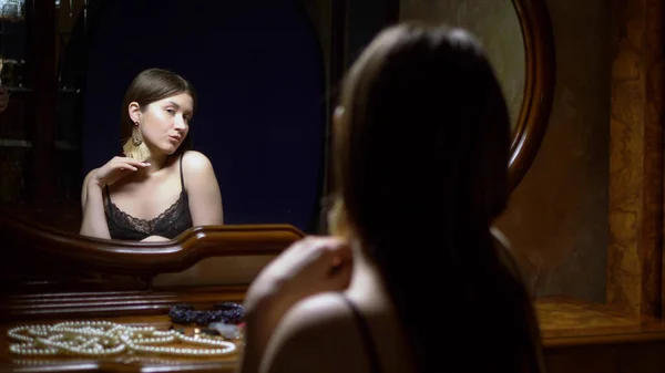 Schöne Mädchen zieht Ohrringe an, sitzt in der Abenddämmerung vor dem Spiegel. — Stockfoto