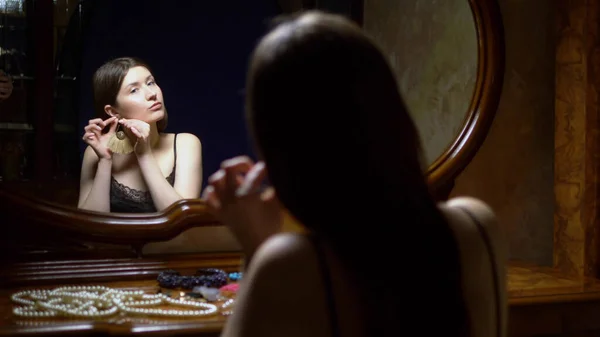 Όμορφο κορίτσι βάζει σκουλαρίκια, κάθεται μπροστά στον καθρέφτη το σούρουπο. — Φωτογραφία Αρχείου