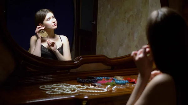 Krásná dívka si nasadí náušnice, sedí před zrcadlem za soumraku. — Stock fotografie