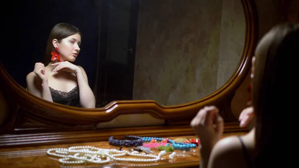 Красивая девушка надевает серьги, сидя перед зеркалом в сумерках . — стоковое фото