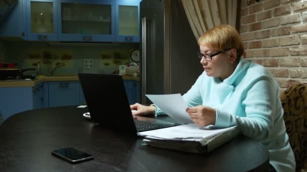 Oudere vrouw die thuis met de documenten werkt met een laptop. — Stockvideo