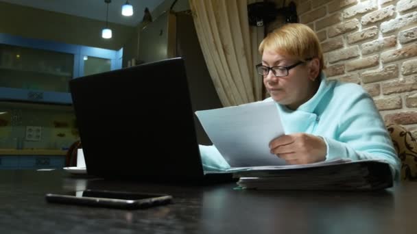 Oudere vrouw die thuis met de documenten werkt met een laptop. — Stockvideo