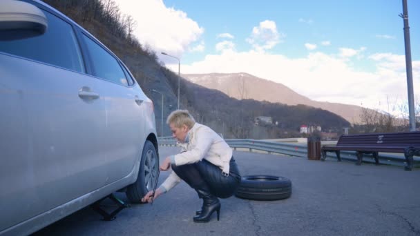Красивая девушка меняет колесо автомобиля, припаркованного на обочине дороги — стоковое видео