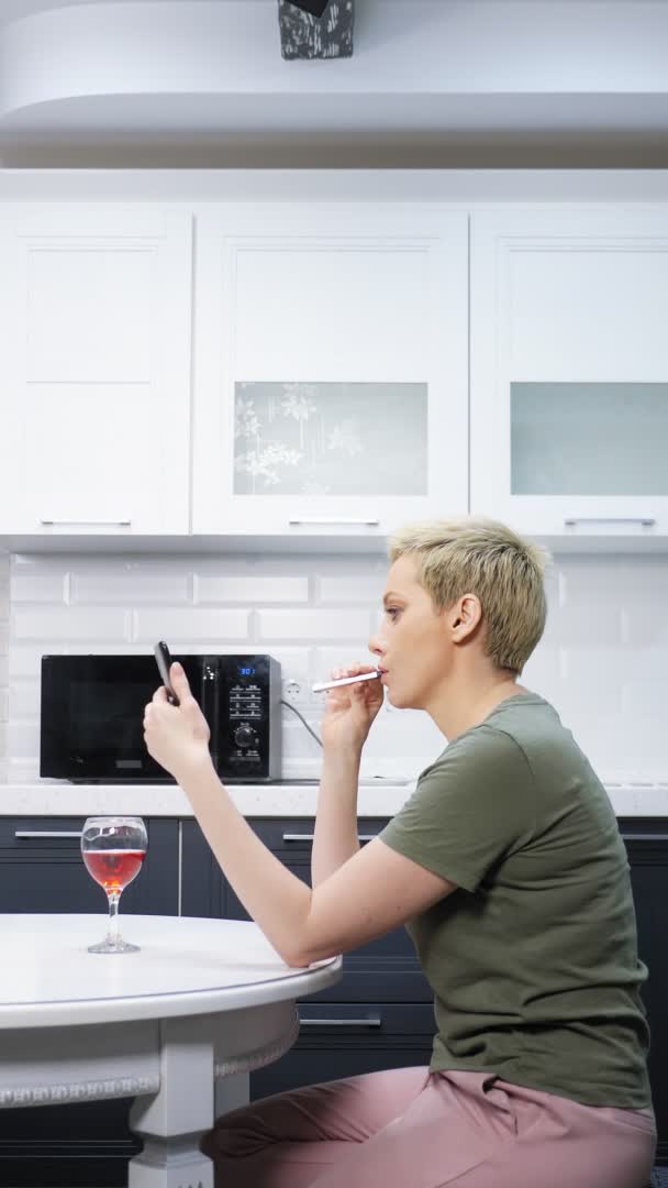 Woman is resting in the kitchen with a glass of wine, e cigar uses a smartphone — Stock Video