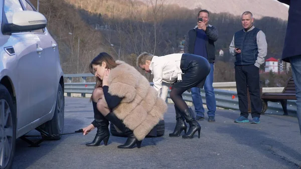 Two girls changing a car wheel on the side, and the men watching from the side — 스톡 사진