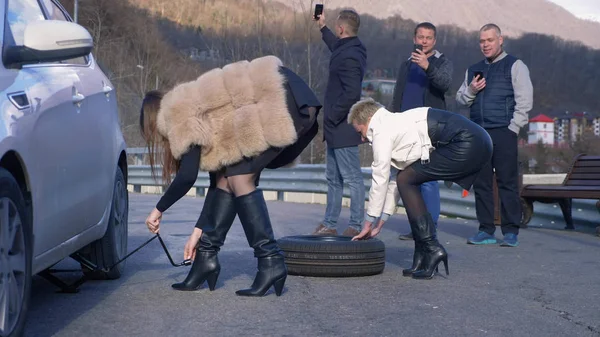 Two girls changing a car wheel on the side, and the men watching from the side — Stock Photo, Image