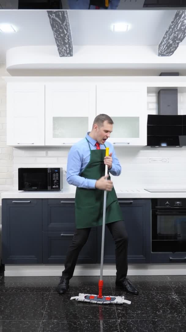 Κάθετη εικόνα. Όμορφος άντρας με γραβάτα και ποδιά σφουγγαρίζει το πάτωμα στην κουζίνα. — Αρχείο Βίντεο