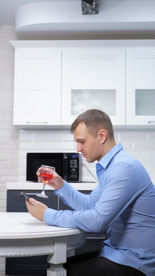 Tiro vertical, el hombre está descansando en la cocina con una copa de vino utiliza un teléfono inteligente — Vídeo de stock
