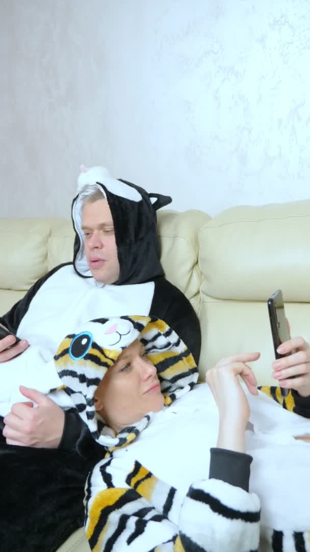 Vertical. um casal em um gato e cachorro kigurumi deitado no sofá e usando smartphone — Vídeo de Stock