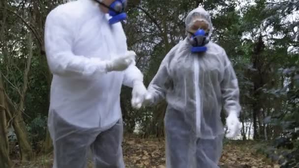 Ein Mann und eine Frau in Schutzanzügen und Atemschutzmasken gehen in den Park — Stockvideo