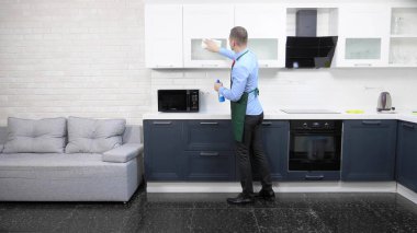 Önlük ve kravat takmış yakışıklı bir adam. Dairedeki mutfağı temizliyor.