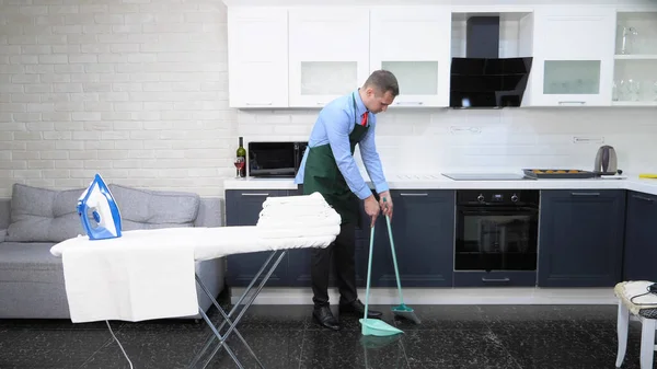 Pohledný muž v kravatě a zástěře zametání podlahy v kuchyni — Stock fotografie