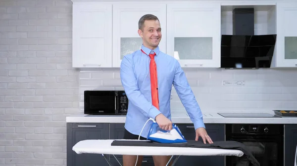 Handsome man in tie ironing pants . the kitchen of his house — Stok fotoğraf
