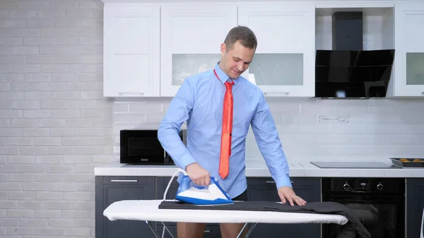 Handsome man in tie ironing pants . the kitchen of his house — Stock fotografie