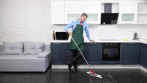 Jóképű férfi nyakkendőben és kötény felmossa a padlót a konyhában — Stock Fotó