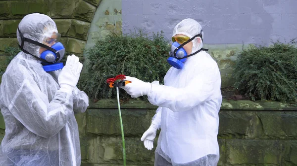 Mężczyzna i kobieta w kombinezonach i respiratorze w parku. Człowiek daje kwiat — Zdjęcie stockowe