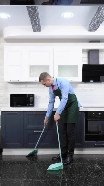 Una imagen vertical. hombre guapo en corbata y delantal barrer el piso de la cocina — Foto de Stock