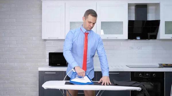 Handsome man in tie ironing pants . the kitchen of his house — ストック写真
