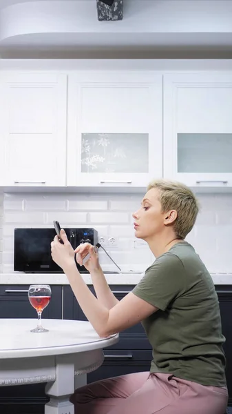 Женщина отдыхает на кухне с бокалом вина, электронная сигара использует смартфон — стоковое фото