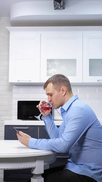 Tiro vertical, el hombre está descansando en la cocina con una copa de vino utiliza un teléfono inteligente —  Fotos de Stock