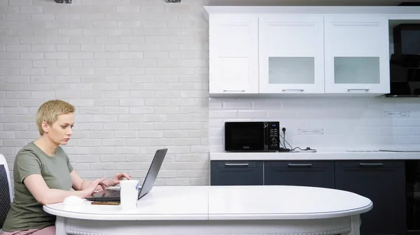 Een vrouw gebruikt een laptop zittend op de keukentafel, ze is gelukkig — Stockfoto