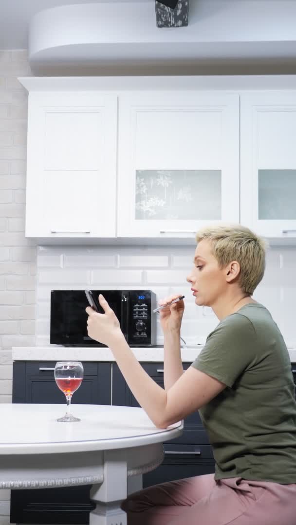 Mujer está descansando en la cocina con una copa de vino, e cigarro utiliza un teléfono inteligente — Vídeos de Stock