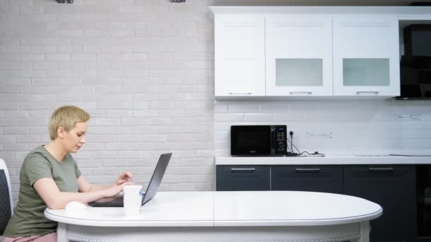 Una donna usa un computer portatile seduto sul tavolo della cucina, lei è felice — Video Stock