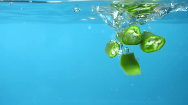 Fette di peperoncino verde tagliate a fette cadendo in acqua. sfondo blu — Video Stock