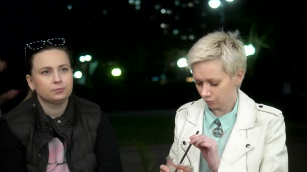 Две женщины разговаривают на улице вечером, пьют коктейли — стоковое видео