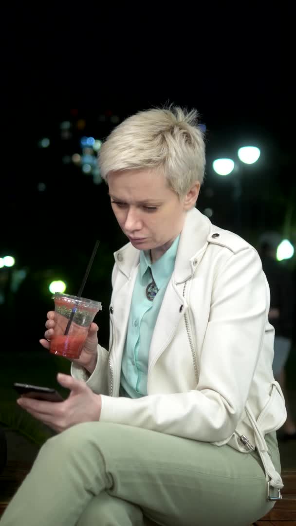 Mujer bebiendo batido al aire libre en la noche. utiliza un teléfono inteligente — Vídeos de Stock