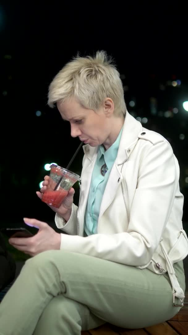 Kvinna dricker smoothie i det fria på kvällen. använder en smartphone — Stockvideo