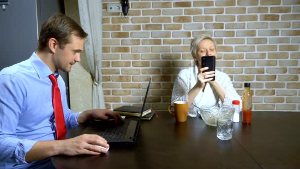 Egy férfi otthon dolgozik a laptopon a konyhában, a feleség eszik. — Stock videók