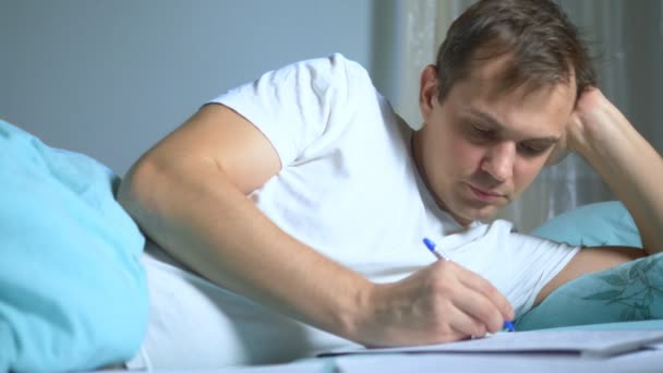 Un uomo scrive su documenti cartacei sdraiato a letto. lavoro a casa . — Video Stock