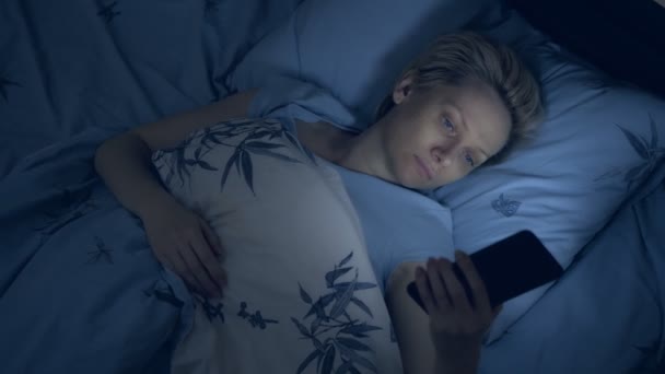 Kvinna som lider av sömnlöshet använder en smartphone när du ligger i sängen i mörkret — Stockvideo