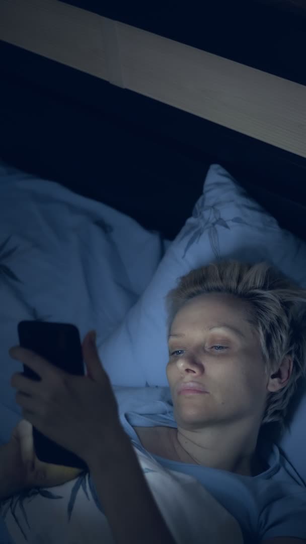 수직으로 요. 불면증으로 고생하는 여자는 침대에 누워 있는 동안 스마트폰을 사용 한다 — 비디오