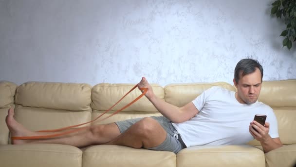 Pria di sofa melakukan olahraga dengan sebuah band elastis dan menggunakan smartphone — Stok Video