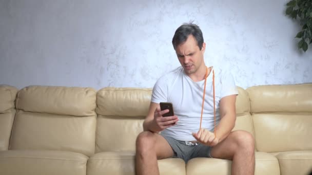 Hombre en el sofá hace deportes con una banda elástica y utiliza un teléfono inteligente — Vídeos de Stock