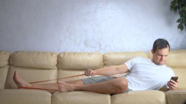 Kanepedeki adam elastik bir bantla spor yapıyor ve akıllı telefon kullanıyor.