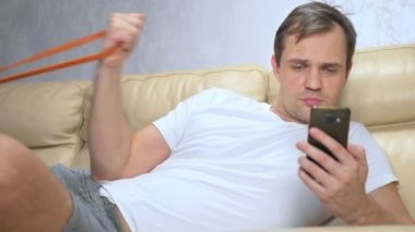 Kanepedeki adam elastik bir bantla spor yapıyor ve akıllı telefon kullanıyor.