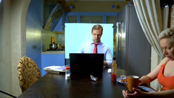 Un hombre tiene una videoconferencia en casa en la cocina su esposa come en la mesa — Vídeos de Stock