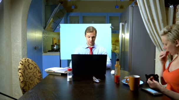Um homem realiza uma videoconferência em casa na cozinha sua esposa come na mesa — Vídeo de Stock