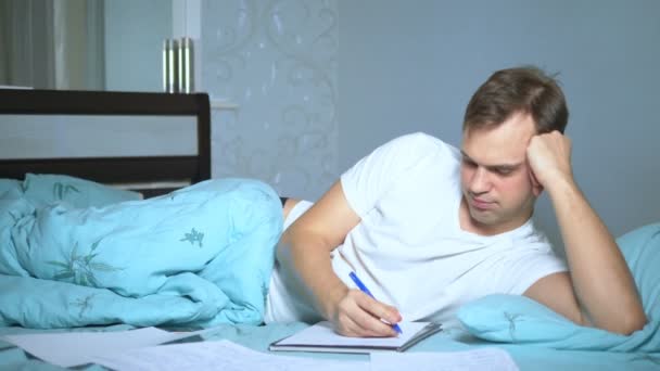 男はベッドに寝そべっている紙の書類に書いてある。家で働いてる. — ストック動画