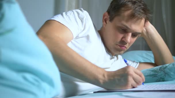Een man schrijft op papieren documenten in bed. thuiswerk. — Stockvideo