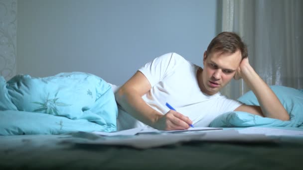 Un uomo scrive su documenti cartacei sdraiato a letto. lavoro a casa . — Video Stock