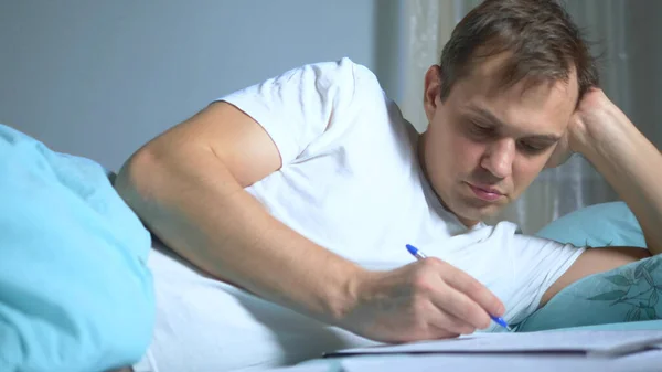 Ein Mann schreibt auf Papierunterlagen, die im Bett liegen. Arbeit zu Hause. — Stockfoto