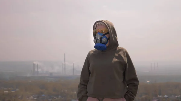 O femeie într-un aparat respirator stă pe fundalul conductelor de fabrică de fumat — Fotografie, imagine de stoc