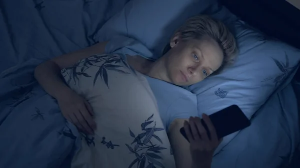 Mulher que sofre de insônia usa um smartphone enquanto deitado na cama no escuro — Fotografia de Stock