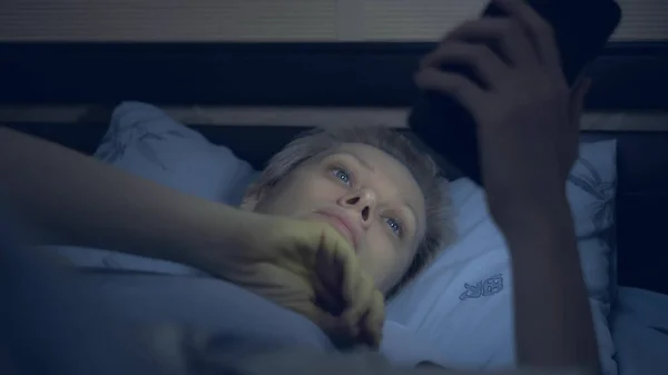 Mulher que sofre de insônia usa um smartphone enquanto deitado na cama no escuro — Fotografia de Stock